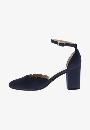 Navy blue suede heeled shoe with scalloped cutouts, an ankle strap, and a chunky block heel. Smooth texture and minimalist design.