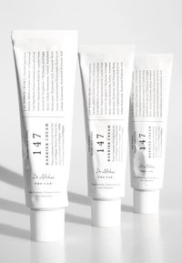 Tre hvite tubecontainere med barrierekrem, med svart tekst som beskriver ingredienser og bruk, med runde hvite lokk.