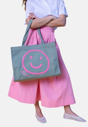 Person trägt ein weißes Hemd und einen rosa Rock und hält eine graue Tragetasche mit einem großen rosa Smiley-Gesicht und dem Text "Fitri".
