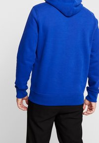 Sudadera azul con un bolsillo tipo canguro en la parte frontal, puños y dobladillo acanalados, y capucha con cordón. La tela parece suave y texturizada.