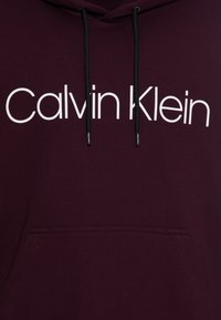 Bordeaux hoodie met een voorzak, voorzien van een witte "Calvin Klein" logo en zwarte koorden. Katoenmix materiaal, zachte textuur.