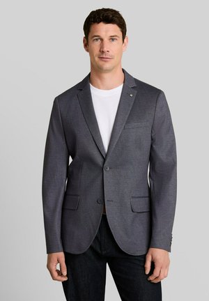 Man draagt een donkergrijze blazer over een wit overhemd en zwarte broek, staand tegen een effen lichtgrijze achtergrond.