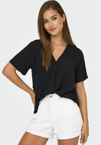 Svart kortärmad V-ringad tröja med strukturerat prickmönster, kombinerad med vita uppvikta denimshorts.