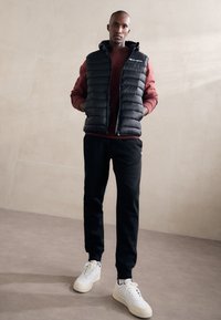 Chaleco acolchado negro sobre una sudadera burdeos, combinado con pantalones de chándal negros y zapatillas blancas. Presenta una capucha y acentos de logo en el chaleco.