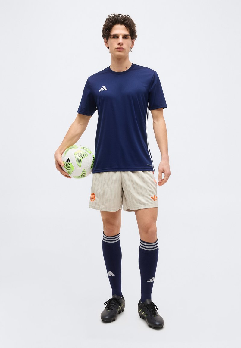 Joven con camiseta deportiva azul marino, pantalones cortos beige a rayas y calcetas azules sosteniendo un balón de fútbol blanco y verde, de pie frente a un fondo liso.