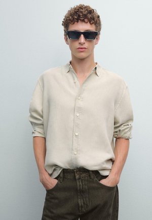 Light beige button-up shirt with rolled sleeves paired with dark green jeans. Model wearing black rectangular sunglasses, with curly hair.