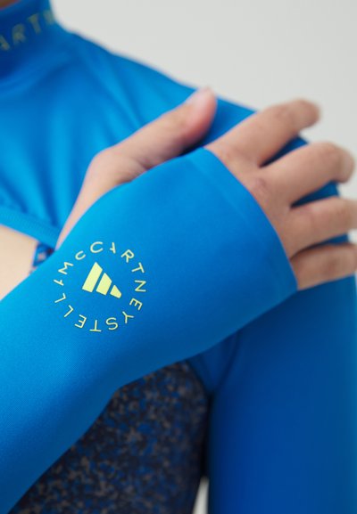 Sininen urheilu paita pitkähihaisilla, jossa on pyöreä keltainen logo hihassa. Suunnassa sileä tekstuuri, istuva muotoilu ja tumma kuvio contrasting kankaassa.