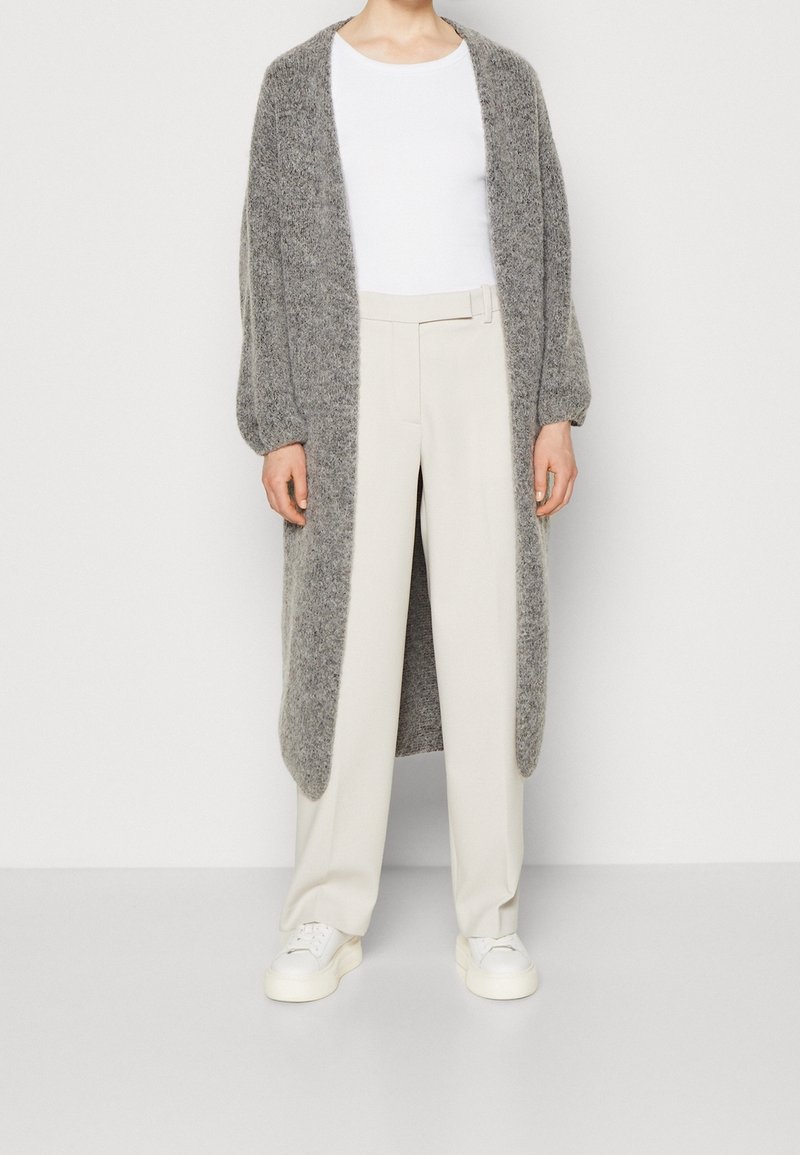 Personne portant un long cardigan en tricot gris, un haut blanc, un pantalon crème taille haute et des baskets blanches sur un fond uni.