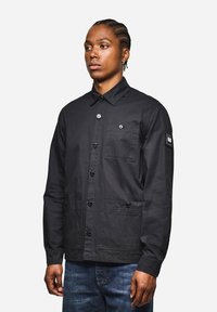 Schwarze Jacke mit Knopfverschluss und Kragen, ausgestattet mit einer aufgesetzten Tasche und zwei Fronttaschen. Hergestellt aus strukturiertem Stoff mit glänzenden Knöpfen.