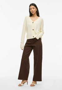 Cardigan côtelé crème avec un col en V et trois grands boutons, associé à un pantalon marron à taille haute et jambes larges, ainsi qu'à des sandales à talons beiges.