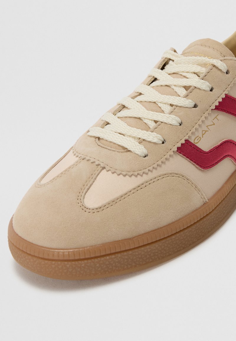 Beżowe sneakersy z zamszowymi i materiałowymi wstawkami, z czerwonymi akcentami, beżowymi sznurówkami i teksturowaną gumową podeszwą. Minimalistyczne detale designu.
