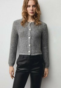 Cardigan gri tricotat cu un model texturat, având un guler rotund și șase nasturi mari. Purtați peste pantaloni negri ajustați.