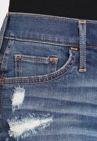 Jeans de denim con un color azul desgastado, que presentan un aspecto envejecido, pequeño bolsillo delantero y costuras naranjas a lo largo de las costuras.