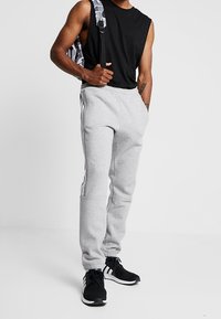 Man som bär en ärmlös svart skjorta, ljusgrå sweatpants med vita ränder, svart och vit sneakers och bär en camouflagemönstrad ryggsäck över ena axeln.