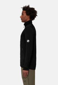 Zwarte zip-up jas met hoge kraag, zijzak met rits en logopatch op de mouw. Het materiaal lijkt glad en aansluitend te zijn.