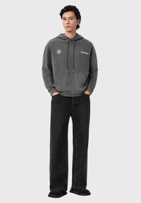 Γκρι φούτερ με κουκούλα, κορδόνια, τσέπη καγκουρό και λογότυπο μπροστά. Συνδυάζεται με μαύρο τζιν φαρδιάς γραμμής και μαύρα παπούτσια. Απλός σχεδιασμός.