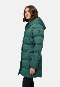 Manteau rembourré teal avec capuche, doté d'une taille cintrée, de poches latérales et d'une pièce en logo ronde sur la manche gauche. Tissu texturé.