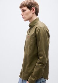 Πουκάμισο σε ελιά πράσινο με κουμπιά, γιακά, μία τσέπη στο στήθος και μικρό κόκκινο έμβλημα. Βαμβακερό ύφασμα με λεία υφή και άνετη εφαρμογή.