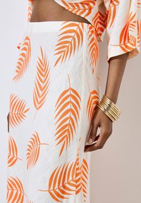 Jupe blanche avec imprimé de feuilles orange, présentant une texture lisse et un tissu léger. Bracelet jonc en or sur une main près de l'ourlet de la jupe.