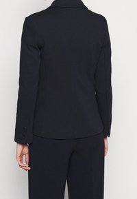 Blazer entallado de un oscuro azul marino con un diseño de un solo botón, cuello con muesca y puños abotonados. Textura suave, forma ajustada y líneas limpias.