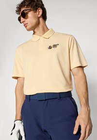 Mann iført beige polo-skjorte med små sushi-tegninger brodert, marineblå bukser, blå belte, hvit golfhanske og brune solbriller.