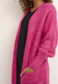 Cardigan lungo e aperto davanti di un vivace colore rosa, realizzato in tessuto a maglia texturizzato. Presenta polsini a coste e tasche laterali. Indossato sopra un top nero.