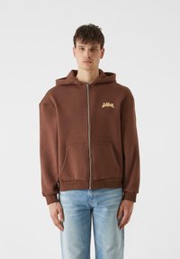 Rudas užtrauktukas džemperis pagamintas iš minkštos medžiagos, su kengūros kišeniu ir gipiūro rankovėmis. Ant krūtinės yra mažas grafikos logotipas.