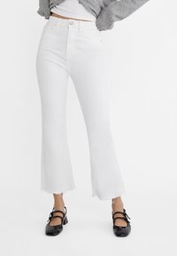Jeans blancs évasés avec un ourlet effiloché, en denim. Associés à des chaussures à talons carrés en cuir verni noir avec deux détails de bride.