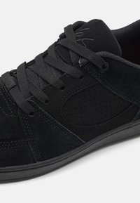 Black suede sneaker with round toe, padded tongue, and laces. Features perforated panel for breathability and rubber sole for grip.