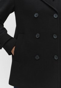 Musta kaksirivinen villakatto, jossa on suuret mustat napit, lovikollar ja sivutaskut. Liukas pinta ja jäsennelty siluetti.