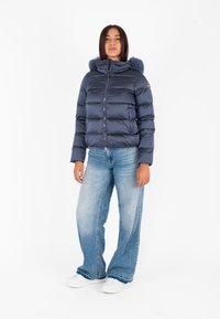 Giacca puffer blu navy con cappuccio foderato in pelliccia, chiusura con zip e baffle orizzontali. Abbinata a jeans a gamba larga blu chiaro e sneakers bianche.