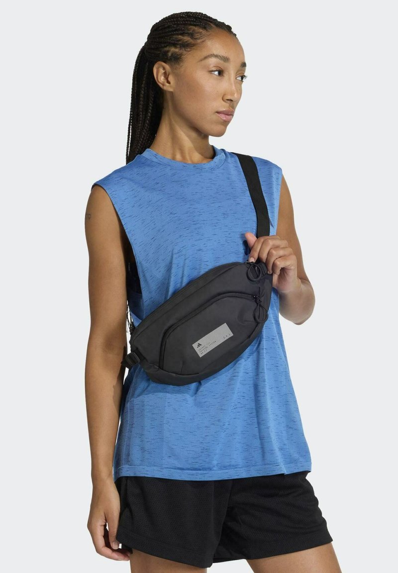 Black fabric waist bag with a smooth texture, zippered compartment, and a reflective logo patch; worn crossbody with adjustable strap.