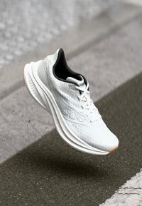 White athletic running shoe with thick sole and black inner lining, captured mid-air above asphalt road near a pedestrian crossing.