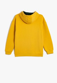 Sudadera amarilla hecha de un tejido suave, con una capucha grande y redondeada y puños acanalados. El interior de la capucha es de color verde oscuro para hacer contraste.