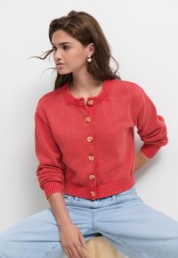 Red knitted cardigan with a round neckline, featuring wooden buttons and ribbed cuffs and hem. Worn over light blue denim.