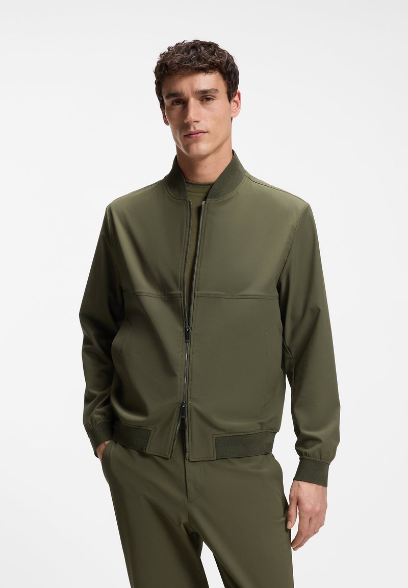 Jeune homme portant une veste bomber vert olive et un pantalon assorti, les mains partiellement dans les poches, debout devant un fond blanc uni.