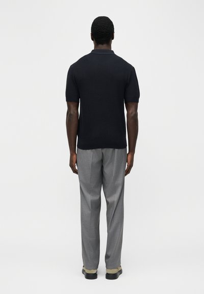 Άνδρας που στέκεται με την πλάτη προς την κάμερα, φορά μαύρο πλεκτό πουκάμισο με κοντά μανίκια, γκρίζο πλισέ παντελόνι και μπεζ παπούτσια, σε απλό φόντο.