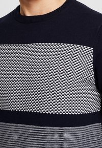 Stickad tröja i marinblått med mönster av vita diamanter över bröstet. Rund halsringning och texturerad detalj vid fållen.