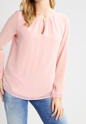Blouse rose clair à manches longues avec un décolleté en forme de trou de serrure, un détail froncé et des poignets élastiqués. Texture lisse, associée à un jean bleu.