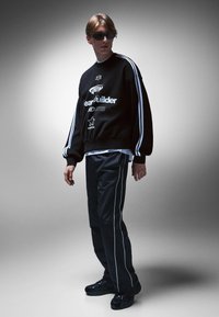 Fekete pulóver fehér csíkokkal, grafikus nyomattal. Fekete nadrág fehér oldalszegéllyel és fekete sportcipő. A modellen napszemüveg van.