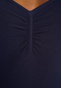 Tmavě modrý pletený top s výstřihem do V a nabranými detaily na přední části, což vytváří texturovaný vzhled.