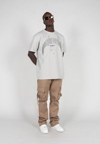 Lys grå grafisk t-skjorte med lilla detaljer, sammen med tan cargo-bukser med flere lommer, og hvite sneakers.