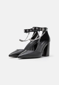 Sorte patentlæder højhælede sko med spids tå, ankelrem med sølvhardware og kædedetaljer, samt en chunky hæl.
