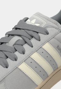 Gros plan sur une basket grise Adidas avec des bandes crème, des lacets gris, une texture en daim, et le logo Adidas sur la languette.