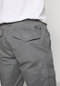 Harmaat cargo-housut rentoutuneella istuvuudella, joissa on useita taskuja, teksturoitu pinta ja merkitty etiketti takana.