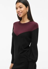 Dzianinowa sukienka w kolorze czarnym i bordowym z zaokrąglonym dekoltem, wzorem w kształcie jodełki na piersi oraz bufkami na długich rękawach. Miękka tekstura.