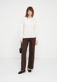 Off-white cable-knit sweater, brown wide-leg pants, black flats, holding a small black bag, standing against a plain white background.