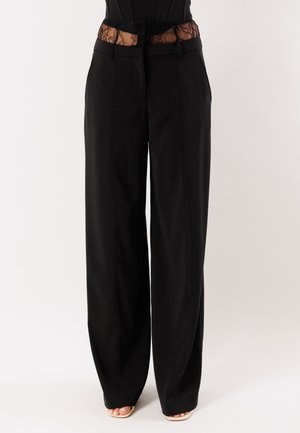 Pantaloni neri a gamba larga indossati con un top corsetto in pizzo nero e tacchi beige aperti, mostrati dalla vita ai piedi su sfondo bianco.