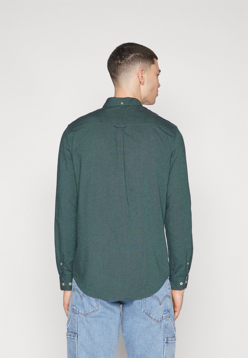 Camisa verde de botonadura con pliegue en la parte posterior, textura suave y mangas largas, combinada con pantalones cargo azul claro.