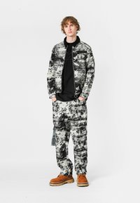 Chaqueta y pantalones con un patrón en blanco y negro, hechos de un material textil; los detalles incluyen una cremallera, puños y unos zapatos naranjas contrastantes.